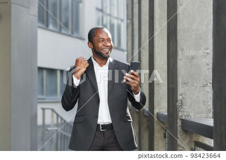 Successful african american boss outside office building using phone businessman in suit celebrating victory, successful achievement, man holding hand up triumph gesture, reading online notification 98423664