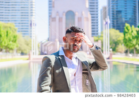 An Indian man wearing a suit standing in a building street and a green park wipes his sweat An Indian man wearing a suit standing in a building street and a green park wipes his sweat 98428403