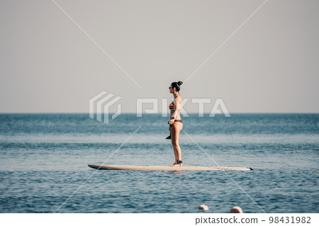 Sea woman sup. Silhouette of happy middle aged tanned woman in rainbow bikini, surfing on SUP board, confident paddling through water surface. Idyllic sunset. Active lifestyle at sea or river. Sea woman sup. Silhouette of happy middle aged tanned woman in rainbow bikini, surfing on SUP board, confident paddling through water surface. Idyllic sunset. Active lifestyle at sea or river. 98431982