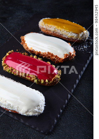Multicolored eclairs on dark background. Custard pastry. Vertical photo. Multicolored eclairs on dark background. Custard pastry. Vertical photo. 98432945