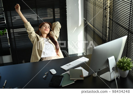 Happy relaxed millennial businesswoman stretching her arms above her head at comfortable office chair Happy relaxed millennial businesswoman stretching her arms above her head at comfortable office chair 98434248