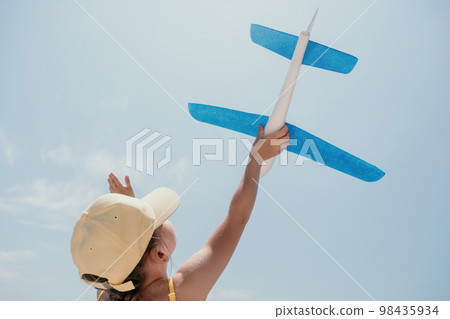 Kid playing with toy airplane. Children dream of travel by plane. Happy child girl has fun in summer vacation by sea and mountains. Outdoors activities at background of blue sky. Lifestyle moment. 98435934