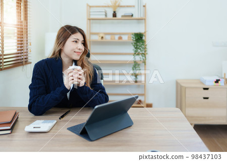 Portrait of an Asian business woman drinking coffee while working with a computer and financial statements documents on her desk. 98437103
