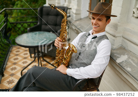 Smiling musician holding saxophone sitting on chair in balcony of his apartment Smiling musician holding saxophone sitting on chair in balcony of his apartment 98437252