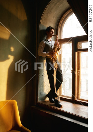Creative young man playing saxophone standing on windowsill 98437265