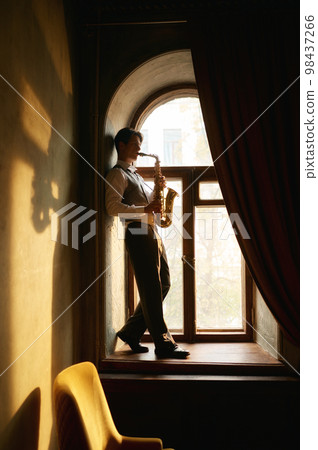 Creative young man playing saxophone standing on windowsill Creative young man playing saxophone standing on windowsill 98437266