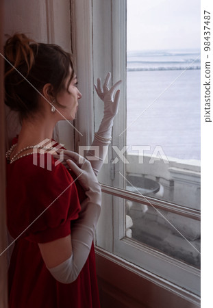Portrait of woman in red dress from 19th century at home looking through the window curiously to the snowy field 98437487