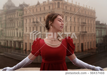 Beautiful young girl in a nineteenth century dress stands on a balcony in front of a beautiful building. 98437488