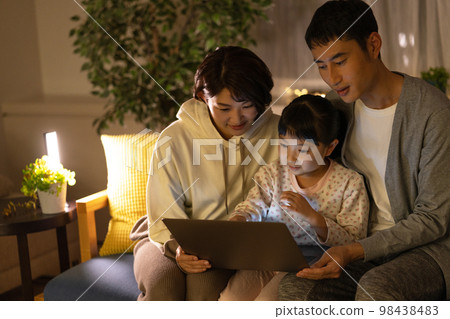 Dad, mom and an elementary school girl sitting side by side on the sofa looking at a laptop at night 98438483