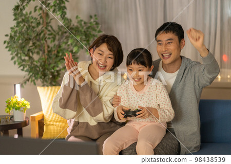 Elementary school girl, dad and mom sitting side by side on the sofa and playing video games at night Elementary school girl, dad and mom sitting side by side on the sofa and playing video games at night 98438539