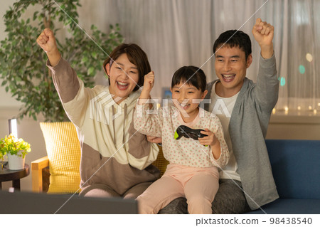 Elementary school girl, dad and mom sitting side by side on the sofa and playing video games at night Elementary school girl, dad and mom sitting side by side on the sofa and playing video games at night 98438540