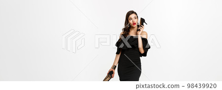 Beauty and fashion concept. Full length of attractive young woman using high heels like mobile phone, standing in black dress against white background 98439920