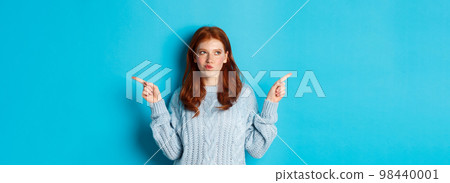 Winter holidays and people concept. Thoughtful redhead girl making decision, pointing fingers sideways, choosing between two ways, standing over blue background 98440001