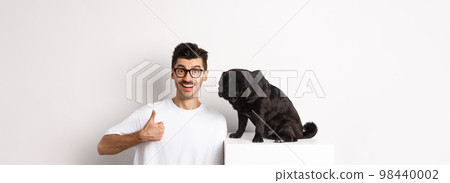Image of happy and satisfied young man sitting near pug dog and showing thumbs-up, smiling and praising good product, white background 98440002
