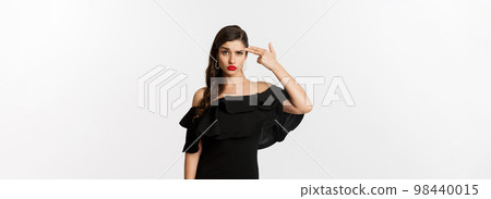 Fashion and beauty. Annoyed young woman in black dress making finger gun gesture near head, shooting herself from irritation, standing over white background 98440015