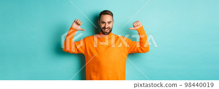 Handsome confident man pointing at himself, looking self-assured, standing in orange sweater against light blue background Handsome confident man pointing at himself, looking self-assured, standing in orange sweater against light blue background 98440019