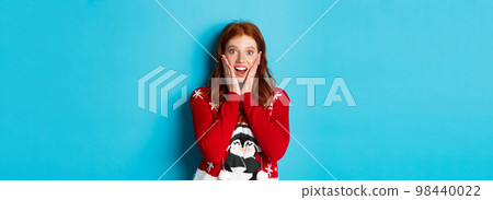 Winter holidays and Christmas Eve concept. Excited redhead girl in xmas sweater looking surprised at camera, standing against blue background 98440022