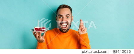 Close-up of happy adult man celebrating birthday, holding bday cake with candle and making wish, standing against turquoise background Close-up of happy adult man celebrating birthday, holding bday cake with candle and making wish, standing against turquoise background 98440036