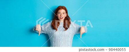 Winter holidays and people concept. Impressed and excited redhead girl, pointing fingers down and showing promo, staring camera amazed, standing over blue background 98440055