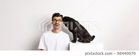 Image of cute black pug dog whispering at owner ear, man looking amazed and smiling, standing over white background Image of cute black pug dog whispering at owner ear, man looking amazed and smiling, standing over white background 98440070