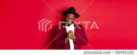 Cunning black man having an idea, smiling devious and rubbing hands, standing against red background and scheming 98440100