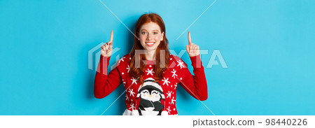 Winter holidays and celebration concept. Cute redhead girl in Christmas sweater, smiling and pointing fingers up at promo logo, standing over blue background 98440226