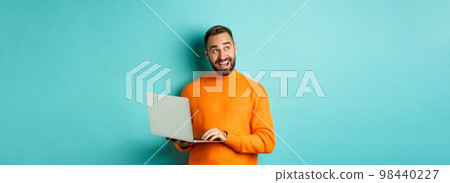 Handsome man doing online shopping, looking up thoughtful while using laptop, standing over light blue background 98440227