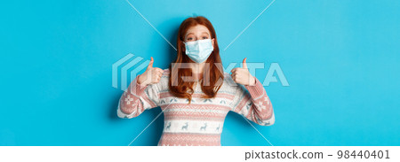 Winter, coronavirus and social distancing concept. Satisfied redhead girl in medical mask and sweater, showing thumbs up in approval, praise something good, blue background 98440401