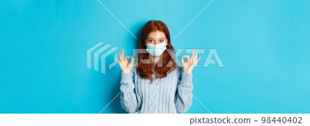 Winter, covid-19 and social distancing concept. Satisfied young redhead woman in face mask showing alright, okay gestures and looking pleased, standing blue background 98440402