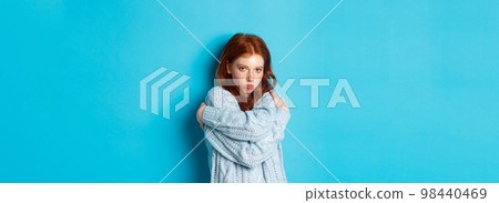Silly and cute redhead girl pucker lips and looking offended, comforting herself by cuddling, embracing body and staring at camera defensive, blue background Silly and cute redhead girl pucker lips and looking offended, comforting herself by cuddling, embracing body and staring at camera defensive, blue background 98440469