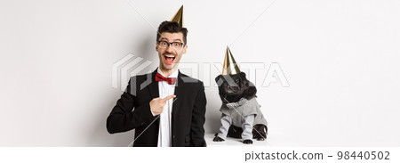 Happy dog owner and black pug wearing birthday party cones, man pointing at pug, standing over white background 98440502