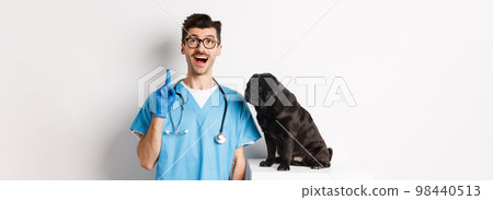 Handsome young doctor at vet clinic pointing finger up and looking amazed, standing near cute black pug dog, white background Handsome young doctor at vet clinic pointing finger up and looking amazed, standing near cute black pug dog, white background 98440513