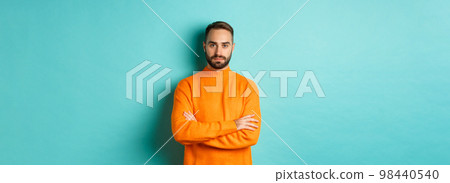 Confident young man looking determined, cross arms on chest, wearing orange winter sweater, standing against turquoise studio background Confident young man looking determined, cross arms on chest, wearing orange winter sweater, standing against turquoise studio background 98440540
