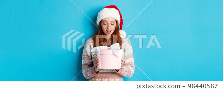 Winter and Christmas Eve concept. Touched and flattered redhead girl looking at box with xmas gift, smiling amazed, standing in santa hat against blue background 98440587