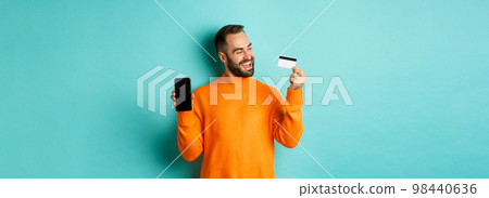 Online shopping. Satisfied man using credit card and showing mobile screen, looking pleased, standing over light blue background 98440636