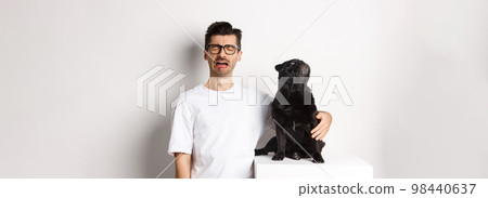 Image of sad crying man hugging his cute black pug, dog looking curious at upper left corner promo logo, standing over white background 98440637