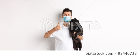 Covid-19, animals and quarantine concept. Young man in medical mask holding cute black pug dog, showing thumb up, like and approve, standing over white background 98440738