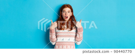 Impressed redhead girl in sweater showing thumbs-up, open mouth fascinated, approve and like product, praising something, standing over blue background 98440770