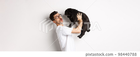 Image of handsome young man loving his pug. Dog owner holding puppy and smiling happy at camera, standing over white background 98440788