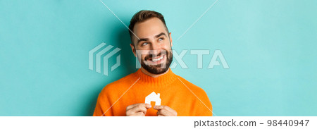 Real estate concept. Happy young man searching for home rent, holding house paper maket and smiling, standing over blue background 98440947
