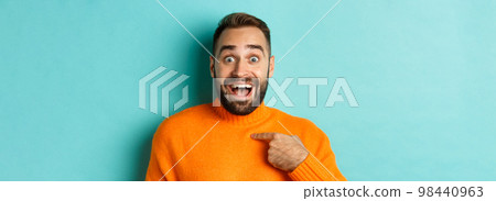 Close-up of happy young man pointing at himself with surprise and excitement, standing over light blue background 98440963