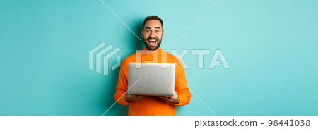 Happy man using laptop and looking excited at camera, standing with computer against light blue background Happy man using laptop and looking excited at camera, standing with computer against light blue background 98441038