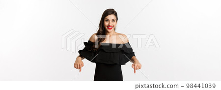 Fashion and beauty. Elegant woman in black dress pointing fingers down, showing promo and smiling, standing over white background Fashion and beauty. Elegant woman in black dress pointing fingers down, showing promo and smiling, standing over white background 98441039
