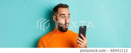 Close-up of caucasian man staring at phone screen with surprised face, wearing orange sweater, light blue background Close-up of caucasian man staring at phone screen with surprised face, wearing orange sweater, light blue background 98441112