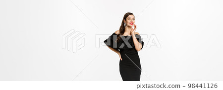 Vertical shot of attractive woman standing in elegant black dress with christmas gifts, smiling happy, standing over white background 98441126
