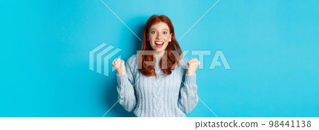 Satisfied redhead girl achieve goal and celebrating, making fist pump gesture and smiling with rejoice, triumphing of win, standing against blue background 98441138