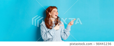 Excited redhead girl in sweater, looking and pointing fingers left, showing promo offer or logo, standing over blue background 98441236