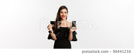 Online shopping concept. Fashionable brunette woman in black dress, showing mobile screen and credit card, smiling pleased, standing over white background 98441238