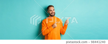 Attractive bearded male model in orange sweater, smiling satisfied and pointing fingers left, standing over turquoise background 98441240