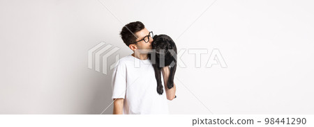 Image of handsome young man kissing his cute black pug, holding dog on shoulder, standing over white background Image of handsome young man kissing his cute black pug, holding dog on shoulder, standing over white background 98441290